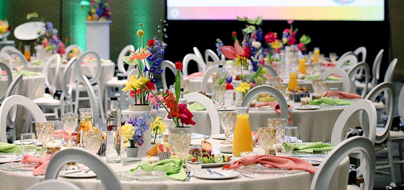  Table settings in a banquet room. Tables include colorful flowers and white chairs and tablecloths.