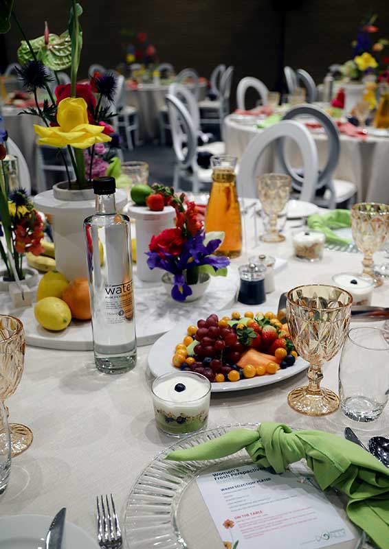 A newly set dinner table within a banquet room. The tables include multiple glasses and colorful fruits and vegetable platters.
